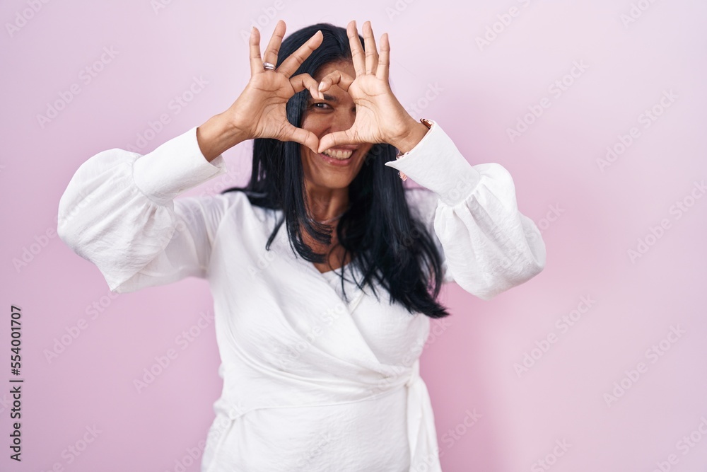 Mature hispanic woman standing over pink background doing heart shape with hand and fingers smiling looking through sign Mature hispanic woman standing over pink background doing heart shape with hand and fingers smiling looking through sign