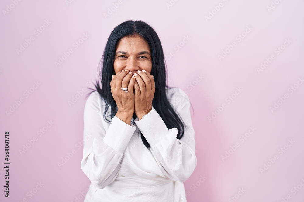 Mature hispanic woman standing over pink background laughing and embarrassed giggle covering mouth with hands, gossip and scandal concept