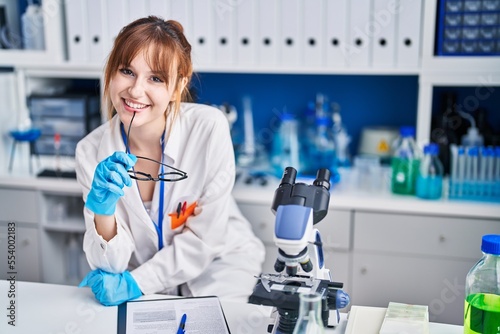 Young woman scientist smiling confident holding glasses at laboratory
