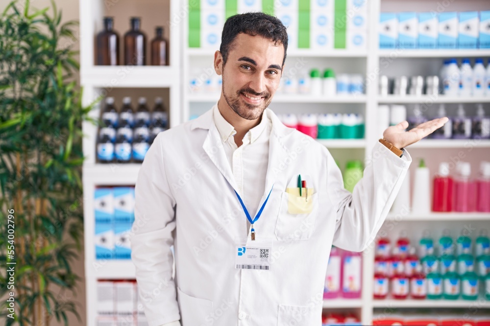 Handsome hispanic man working at pharmacy drugstore smiling cheerful presenting and pointing with palm of hand looking at the camera.
