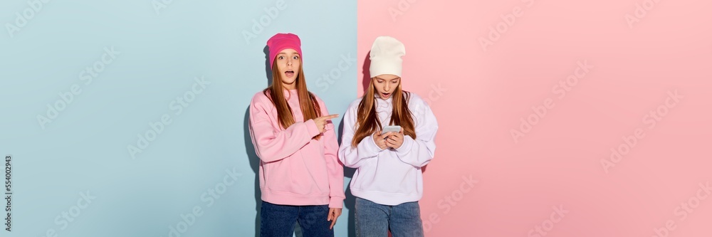Two surprised teen girls in trendy warm clothes and hats using phone isolated over pink-blue background. Youth fashion, beauty, emotions, friendship and ad Two surprised teen girls in trendy warm clothes and hats using phone isolated over pink-blue background. Youth fashion, beauty, emotions, friendship and ad