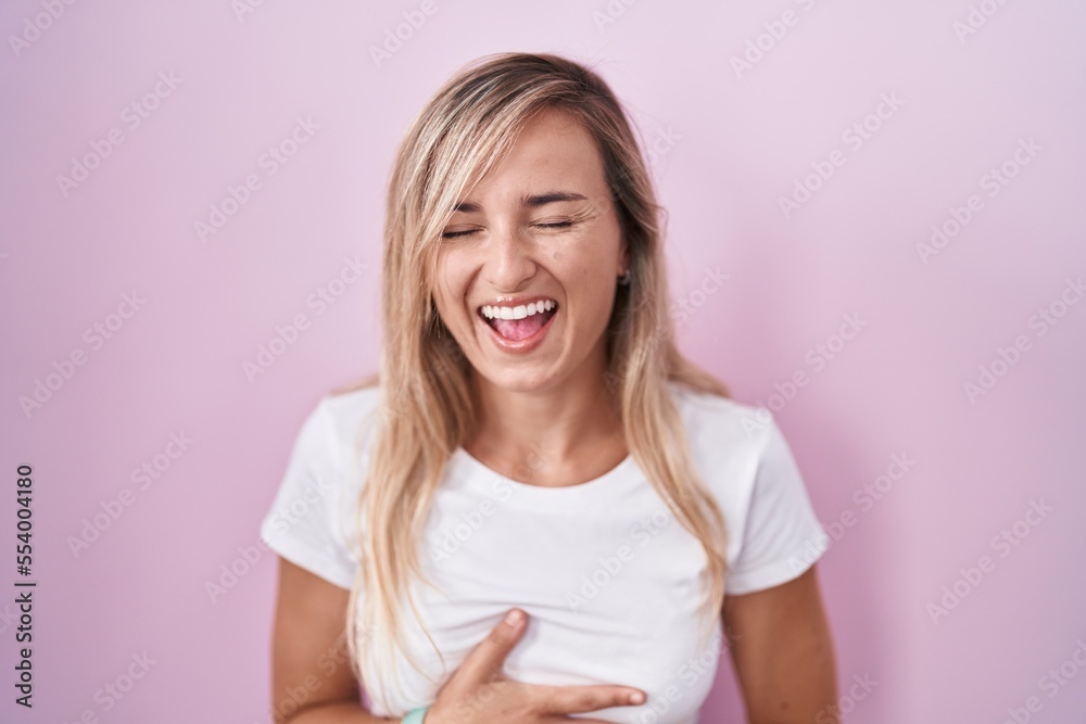 Young blonde woman standing over pink background smiling and laughing hard out loud because funny crazy joke with hands on body. Young blonde woman standing over pink background smiling and laughing hard out loud because funny crazy joke with hands on body.