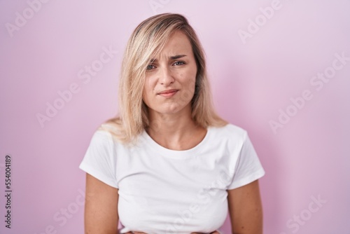 Young blonde woman standing over pink background with hand on stomach because indigestion, painful illness feeling unwell. ache concept.