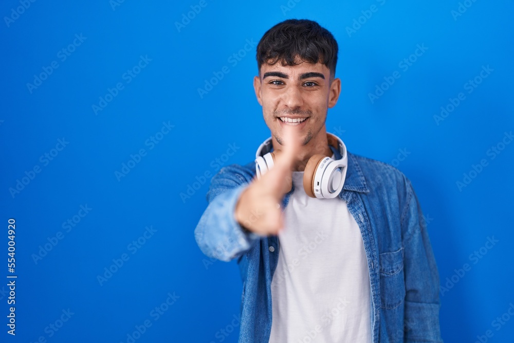 Young hispanic man standing over blue background smiling friendly offering handshake as greeting and welcoming. successful business. Young hispanic man standing over blue background smiling friendly offering handshake as greeting and welcoming. successful business.