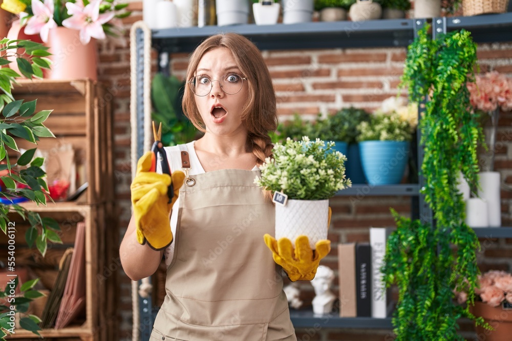 Young caucasian woman working at florist shop holding plant in shock face, looking skeptical and sarcastic, surprised with open mouth Young caucasian woman working at florist shop holding plant in shock face, looking skeptical and sarcastic, surprised with open mouth