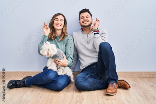 Young hispanic couple sitting on the floor with dog gesturing finger crossed smiling with hope and eyes closed. luck and superstitious concept.
