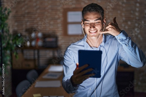 Canvas Print Handsome hispanic man working at the office at night smiling doing phone gesture with hand and fingers like talking on the telephone