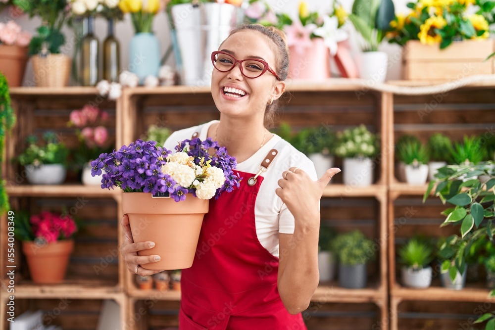 Young hispanic woman working at florist shop holding plant pointing thumb up to the side smiling happy with open mouth