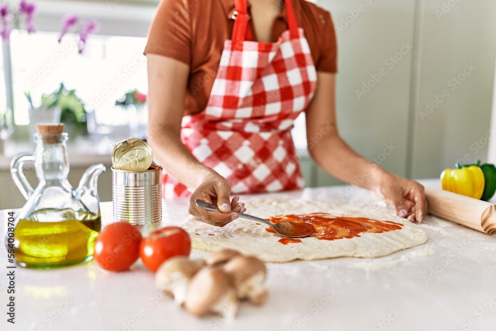 Young beautiful hispanic woman putting tomato sauce on dough pizza at the kitchen Young beautiful hispanic woman putting tomato sauce on dough pizza at the kitchen