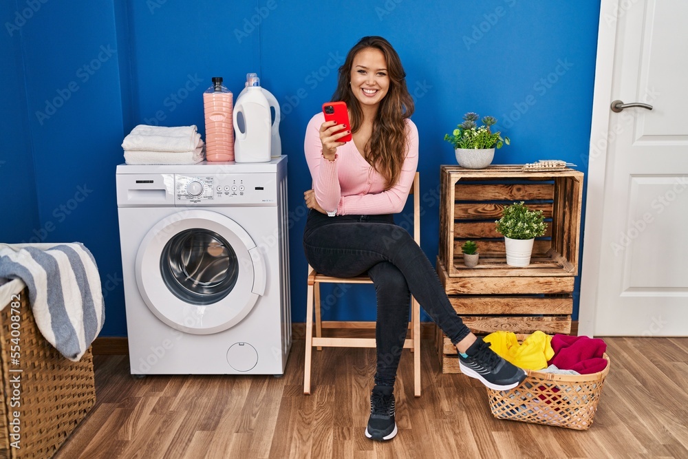 Young hispanic woman sitting waiting for laundry using smartphone with a happy and cool smile on face. lucky person. Young hispanic woman sitting waiting for laundry using smartphone with a happy and cool smile on face. lucky person.