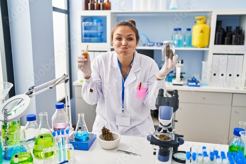Young hispanic woman doing weed oil extraction at laboratory puffing cheeks with funny face. mouth inflated with air, catching air.