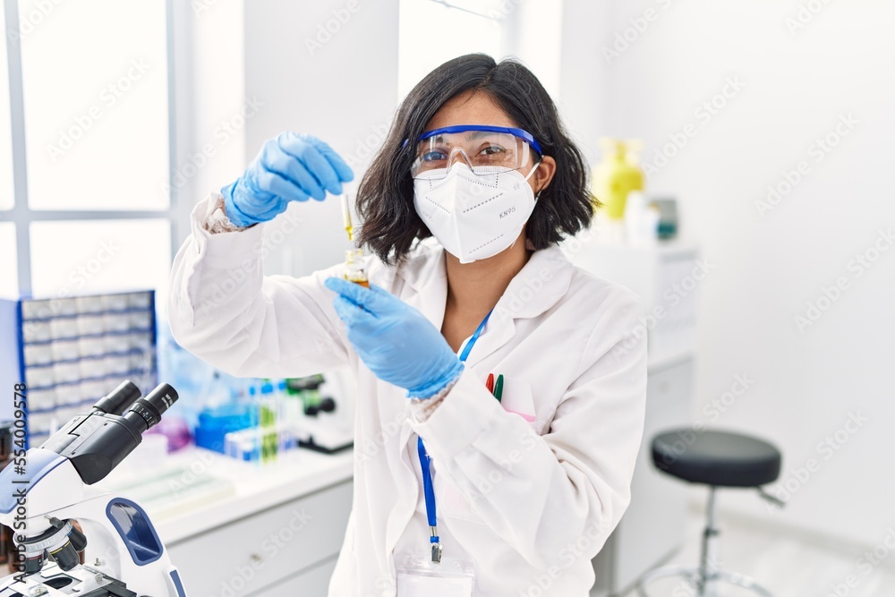 Young latin woman wearing scientist uniform and medical mask using pipette at laboratory Young latin woman wearing scientist uniform and medical mask using pipette at laboratory