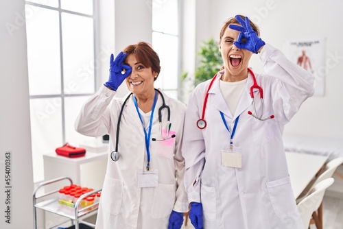 Two women wearing doctor uniform and stethoscope smiling happy doing ok sign with hand on eye looking through fingers