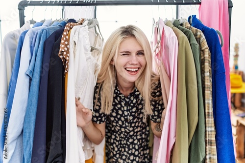 Young blonde woman searching clothes on clothing rack winking looking at the camera with sexy expression, cheerful and happy face.