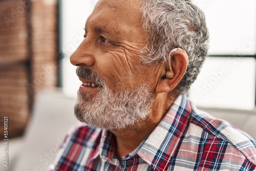 Senior grey-haired man using hearing aid sitting on sofa at home