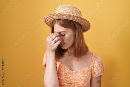 Young redhead woman standing over yellow background wearing summer hat tired rubbing nose and eyes feeling fatigue and headache. stress and frustration concept.