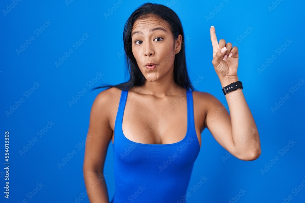 Hispanic woman standing over blue background pointing finger up with successful idea. exited and happy. number one.