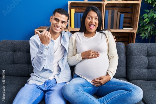 Young hispanic couple expecting a baby sitting on the sofa at home pointing thumb up to the side smiling happy with open mouth