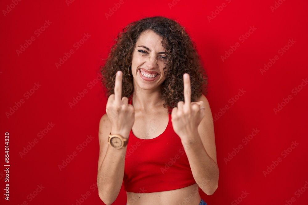 Hispanic woman with curly hair standing over red background showing middle finger doing fuck you bad expression, provocation and rude attitude. screaming excited Hispanic woman with curly hair standing over red background showing middle finger doing fuck you bad expression, provocation and rude attitude. screaming excited
