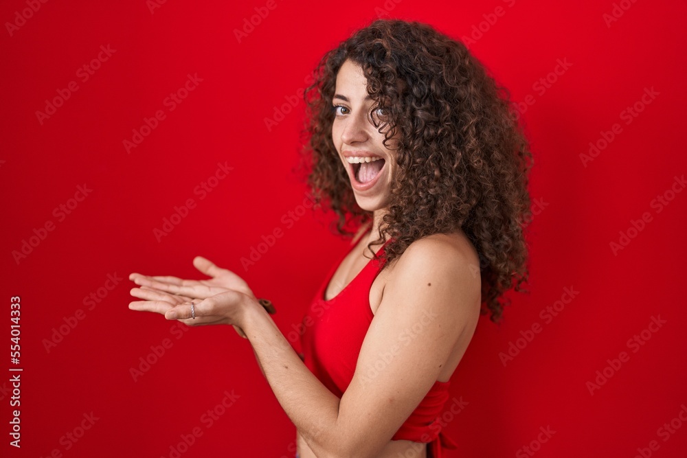 Hispanic woman with curly hair standing over red background pointing aside with hands open palms showing copy space, presenting advertisement smiling excited happy Hispanic woman with curly hair standing over red background pointing aside with hands open palms showing copy space, presenting advertisement smiling excited happy