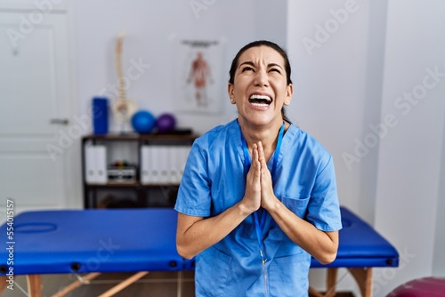 Obraz na plátně Young hispanic woman wearing physiotherapist uniform standing at clinic begging and praying with hands together with hope expression on face very emotional and worried