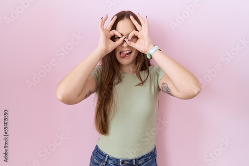Beautiful brunette woman standing over pink background doing ok gesture like binoculars sticking tongue out, eyes looking through fingers. crazy expression. © Krakenimages.com