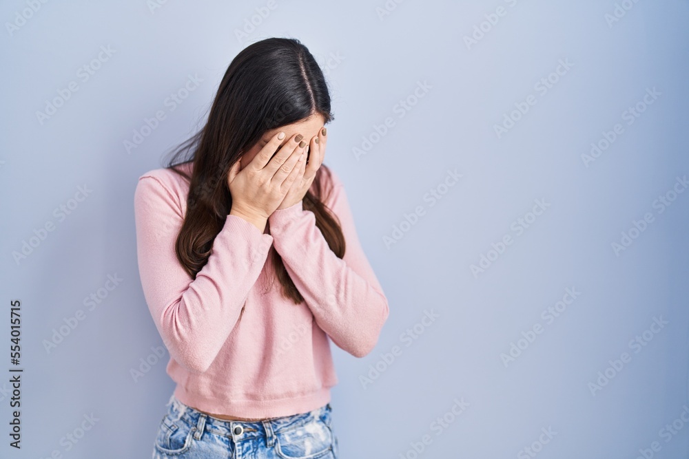 Young brunette woman standing over blue background with sad expression covering face with hands while crying. depression concept. Young brunette woman standing over blue background with sad expression covering face with hands while crying. depression concept.