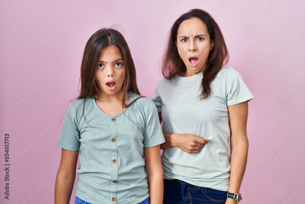 Young mother and daughter standing over pink background in shock face, looking skeptical and sarcastic, surprised with open mouth Young mother and daughter standing over pink background in shock face, looking skeptical and sarcastic, surprised with open mouth