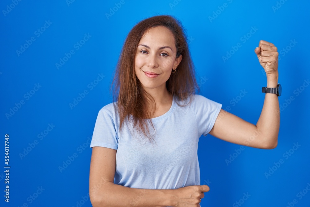 Brunette woman standing over blue background strong person showing arm muscle, confident and proud of power