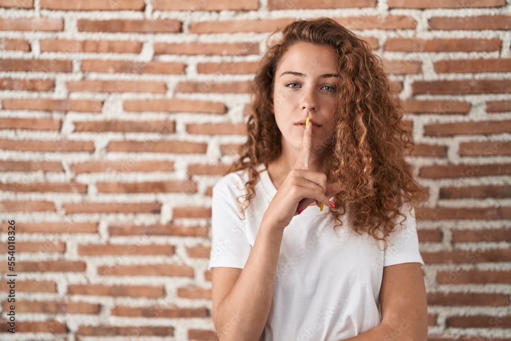 Young caucasian woman standing over bricks wall background asking to be quiet with finger on lips. silence and secret concept.