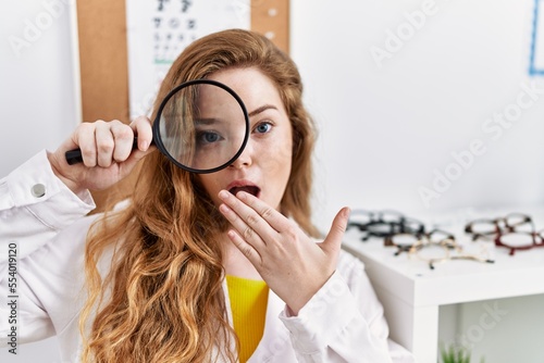 Young caucasian optician woman holding magnifying glass at the clinic covering mouth with hand, shocked and afraid for mistake. surprised expression