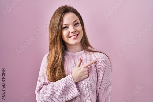 Young caucasian woman standing over pink background cheerful with a smile on face pointing with hand and finger up to the side with happy and natural expression