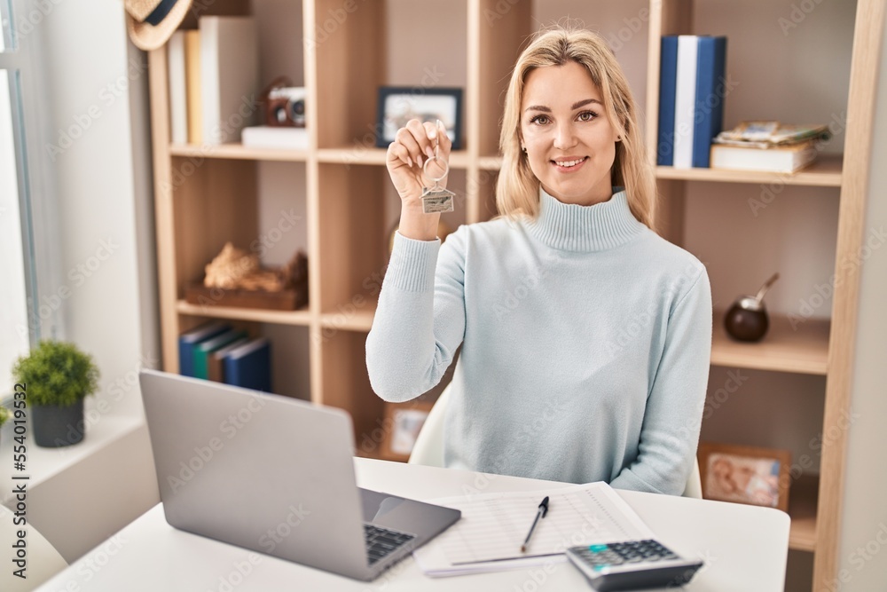 Young caucasian woman holding keys of new home looking positive and happy standing and smiling with a confident smile showing teeth Young caucasian woman holding keys of new home looking positive and happy standing and smiling with a confident smile showing teeth