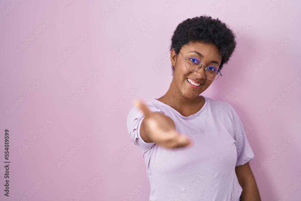 Young african american woman standing over pink background smiling friendly offering handshake as greeting and welcoming. successful business. Young african american woman standing over pink background smiling friendly offering handshake as greeting and welcoming. successful business.