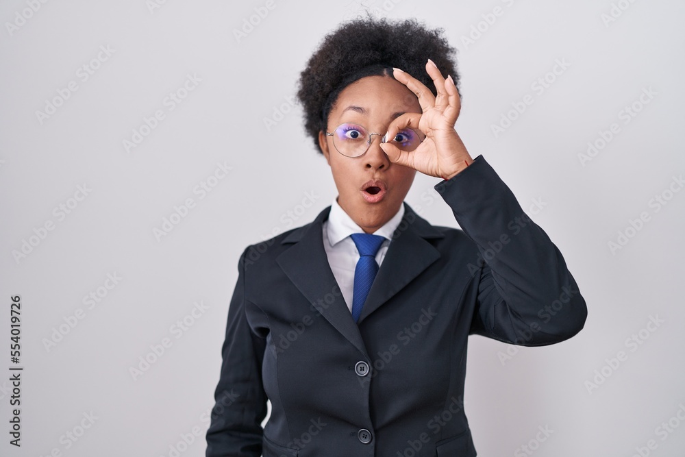 Beautiful african woman with curly hair wearing business jacket and glasses doing ok gesture shocked with surprised face, eye looking through fingers. unbelieving expression.