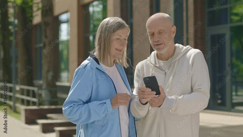 Senior couple is standing on the street after the training and listens to music, podcast or video chat with smartphone. Sport, technology, communication and healthy lifestyle concept Senior couple is standing on the street after the training and listens to music, podcast or video chat with smartphone. Sport, technology, communication and healthy lifestyle concept