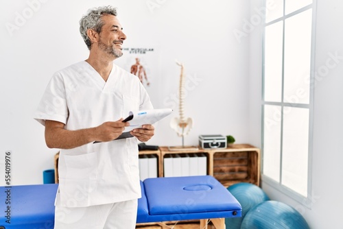 Middle age grey-haired man pysiotherapist smiling confident reading document at rehab clinic