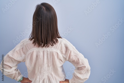 Middle age hispanic woman standing over blue background standing backwards looking away with arms on body
