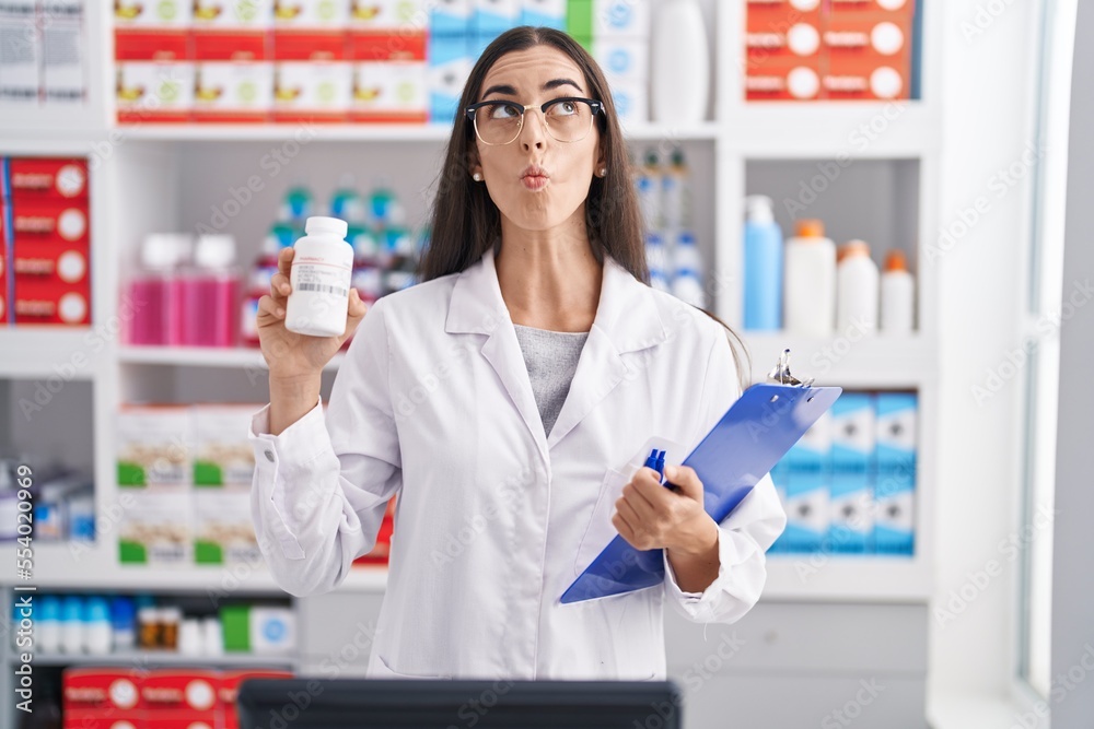 Young brunette woman working at pharmacy drugstore holding pills making fish face with mouth and squinting eyes, crazy and comical.