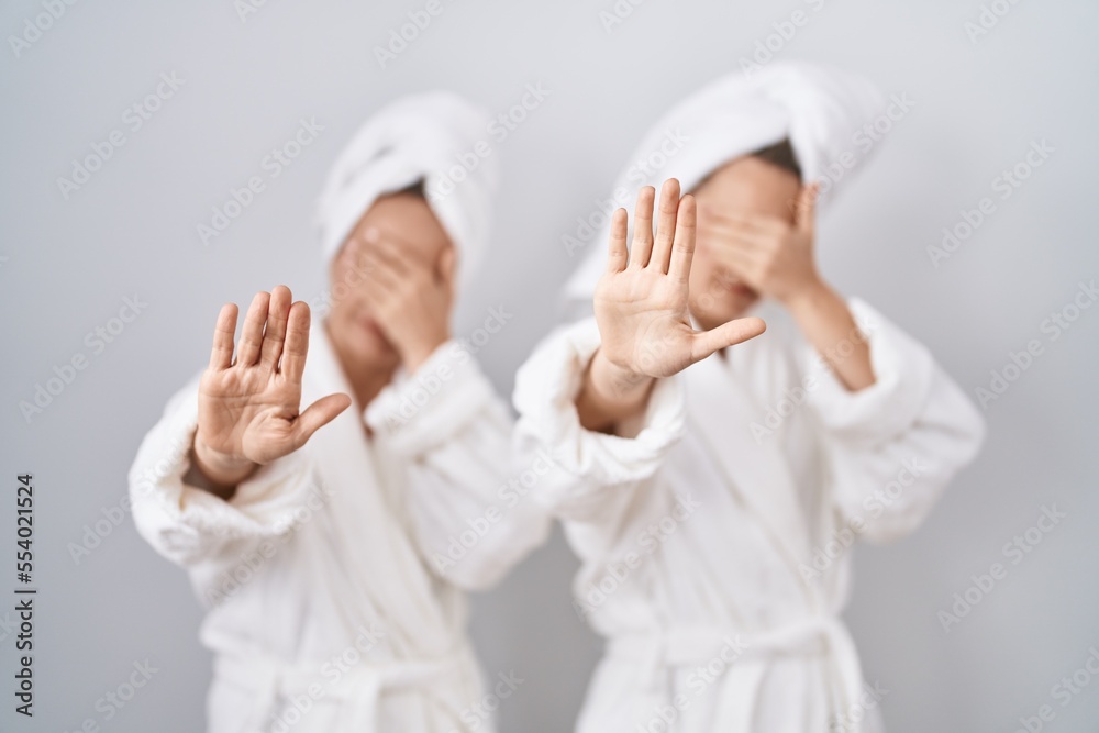 Middle age woman and daughter wearing white bathrobe and towel covering eyes with hands and doing stop gesture with sad and fear expression. embarrassed and negative concept. Middle age woman and daughter wearing white bathrobe and towel covering eyes with hands and doing stop gesture with sad and fear expression. embarrassed and negative concept.