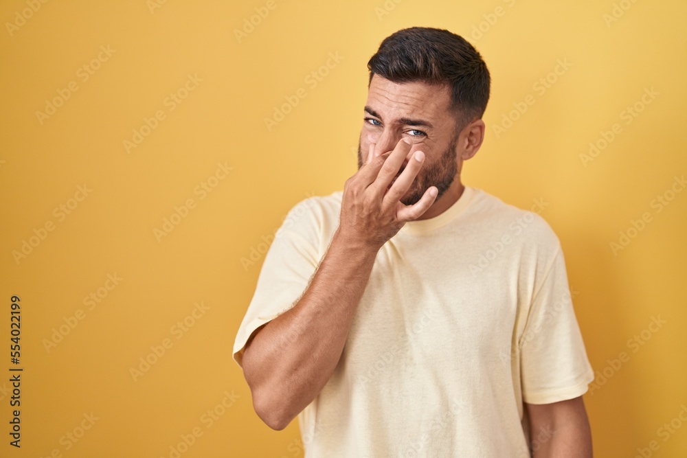 Handsome hispanic man standing over yellow background smelling something stinky and disgusting, intolerable smell, holding breath with fingers on nose. bad smell Handsome hispanic man standing over yellow background smelling something stinky and disgusting, intolerable smell, holding breath with fingers on nose. bad smell