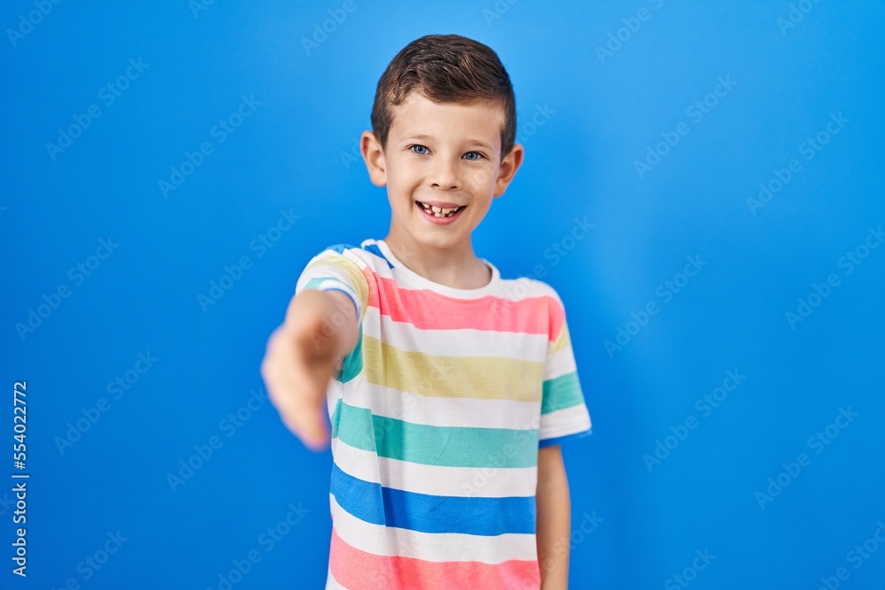 Young caucasian kid standing over blue background smiling friendly offering handshake as greeting and welcoming. successful business. Young caucasian kid standing over blue background smiling friendly offering handshake as greeting and welcoming. successful business.