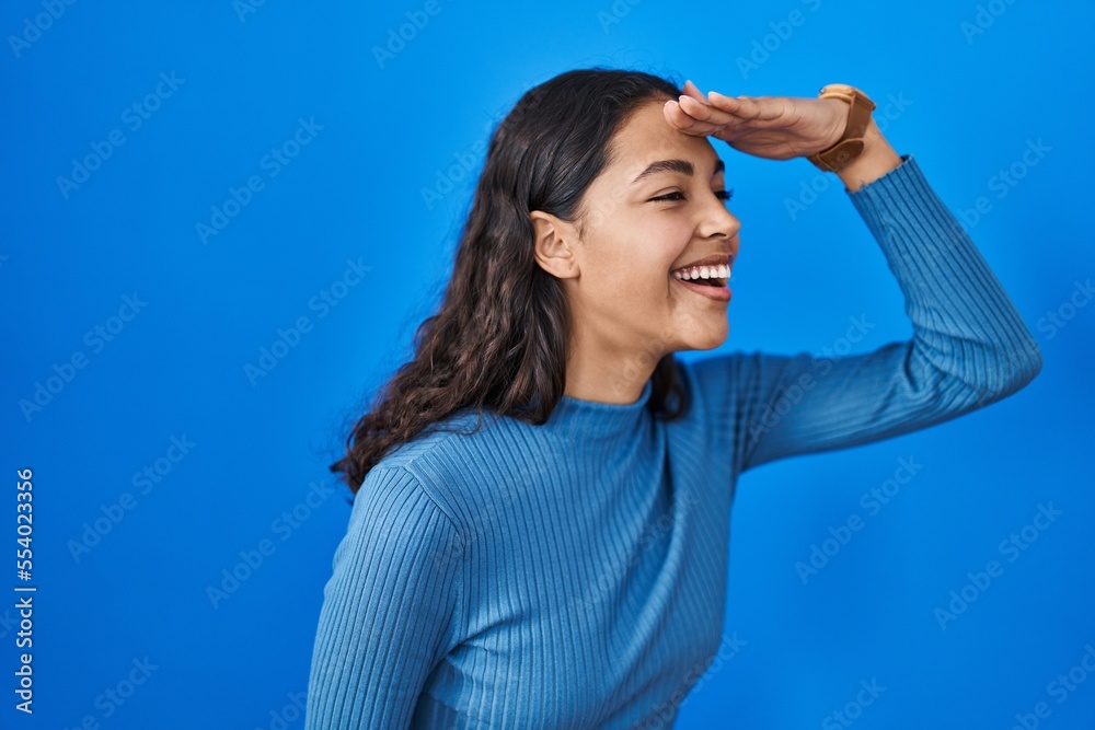 Young brazilian woman standing over blue isolated background very happy and smiling looking far away with hand over head. searching concept. Young brazilian woman standing over blue isolated background very happy and smiling looking far away with hand over head. searching concept.