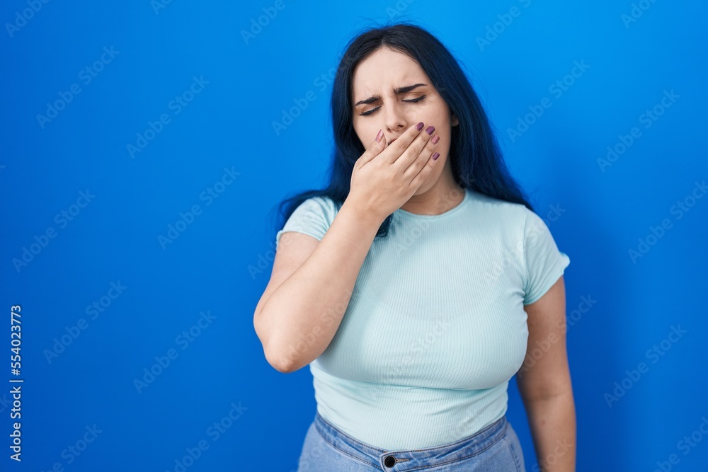 Young modern girl with blue hair standing over blue background bored yawning tired covering mouth with hand. restless and sleepiness. Young modern girl with blue hair standing over blue background bored yawning tired covering mouth with hand. restless and sleepiness.