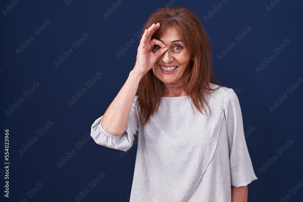 Middle age woman standing over blue background doing ok gesture with hand smiling, eye looking through fingers with happy face. Middle age woman standing over blue background doing ok gesture with hand smiling, eye looking through fingers with happy face.