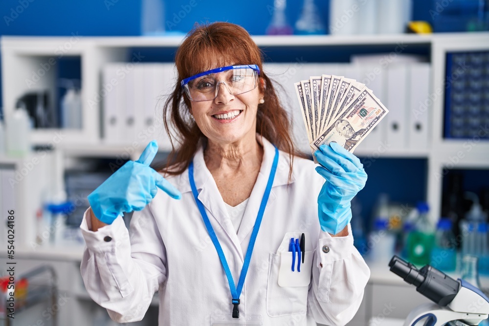 Middle age hispanic woman working at scientist laboratory holding dollars pointing finger to one self smiling happy and proud Middle age hispanic woman working at scientist laboratory holding dollars pointing finger to one self smiling happy and proud