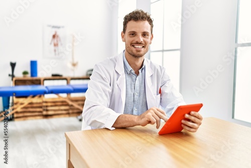 Young hispanic man wearing physiotherapist uniform using touchpad at clinic