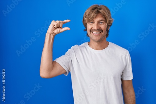 Middle age man standing over blue background smiling and confident gesturing with hand doing small size sign with fingers looking and the camera. measure concept.