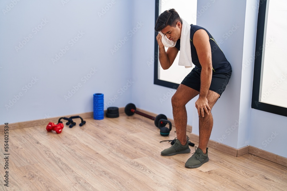 Young hispanic man leaning on wall relaxing using towel at sport center
