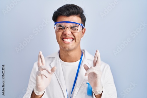 Hispanic man working as scientist gesturing finger crossed smiling with hope and eyes closed. luck and superstitious concept.
